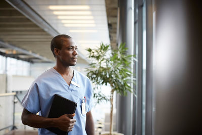 Thoughtful mid adult male nurse holding digital tablet while looking through window in corridor at hospital