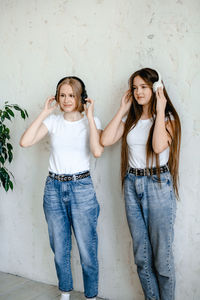 Two teenage girls stand near a white wall in funny sunglasses. . headphones hang on their necks