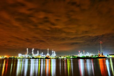 Illuminated factory by sea against sky at night