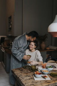 Man kissing smiling non-binary person while having breakfast at home