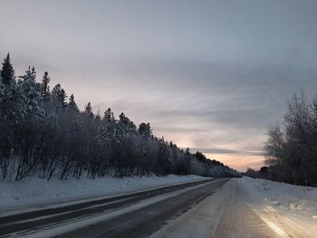 Road amidst snow covered trees against sky during sunset