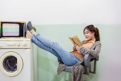 Full length of young woman reading book while relaxing against wall on chair