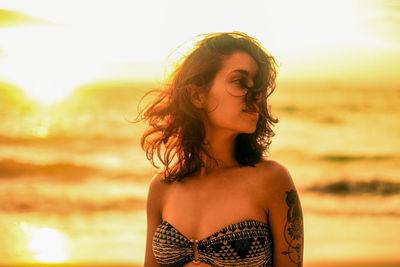 Thoughtful young woman with tousled hair standing at beach during sunset