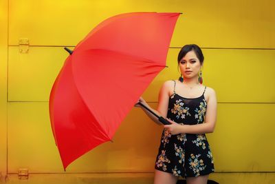 Portrait of young woman with red umbrella standing against yellow wall
