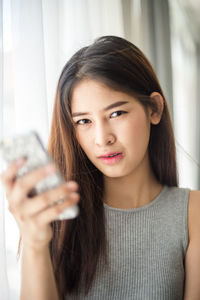 Close-up portrait of young woman using phone by curtain at home