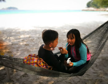 Siblings eating while sitting on hammock at beach