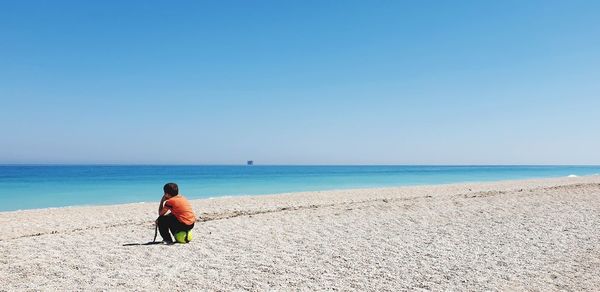 Boy sitting at sandy beach against clear sky