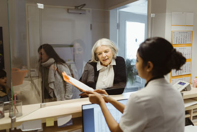 Senior female patient understanding form filling procedure by receptionist through transparent shield in clinic