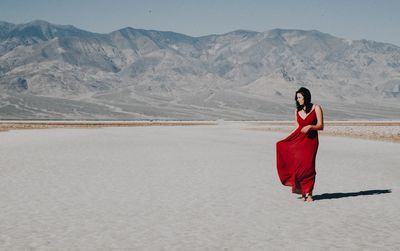 Full length of young woman in red dress standing on sand dune against mountain during sunny day