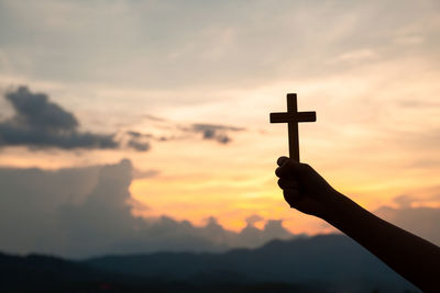 Silhouette person holding cross against sky during sunset