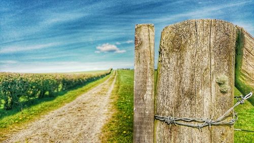 Close-up of wooden fence on field by dirt road against sky during sunny day