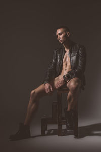 Young man looking away while sitting on chair against black background