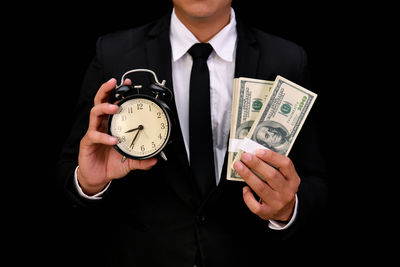 Midsection of businessman holding currency and clock against black background