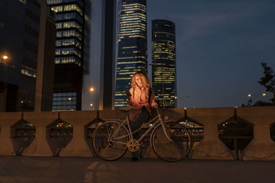 Young woman with bicycle using mobile phone in modern city at night