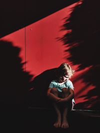 Sad boy sitting against red wall with sunlight