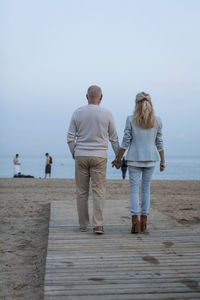 Spain, barcelona, rear view of senior couple walking hand in hand on the beach at dusk