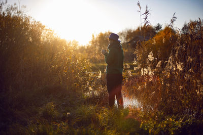 Woman in knitted sweater and blue hat stands bushes sunset and drinks tea from an iron thermos mug