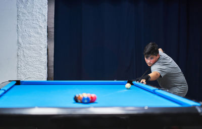 Serious boy in casual wear with cue stick preparing to hit ball while playing pool game at blue billiard table