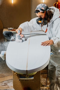 Male shaper in protective respirator and costume using electric planer and polishing surface of surfboard in workshop