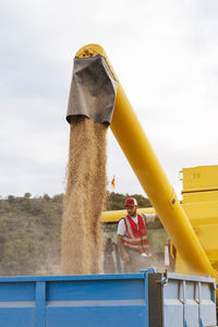 Industrial combine harvester unloading dried yellow grains into trailer in agricultural field with green trees on summer day in countryside