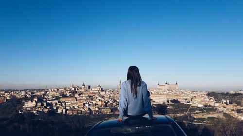 Rear view of woman looking at cityscape while sitting on car roof against clear blue sky