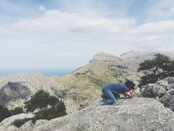 Man photographing with camera on rocks at mountain against cloudy sky