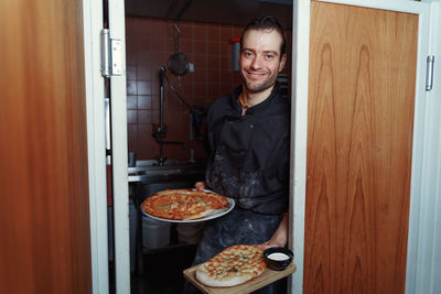 Smiling male chef looking at camera and showing a pizzas against kitchen 