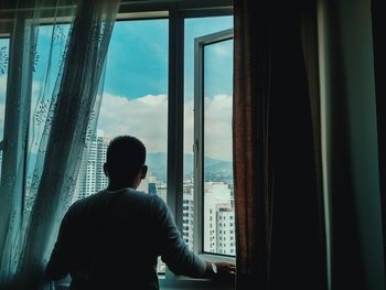 Rear view of man standing amidst curtains at window