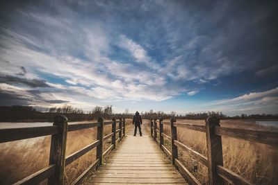 Rear view of man standing on boardwalk against cloudy sky