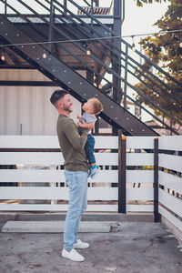 Dad laughing while holding boy in front of white fence and black steps