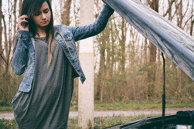 Young woman talking on mobile phone while looking at engine of car