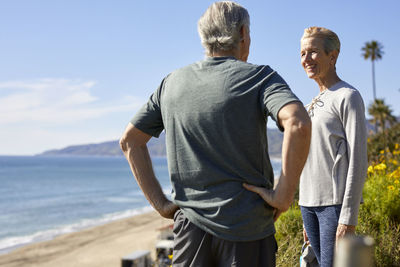 Happy senior couple talking on cliff at beach against sky during sunny day