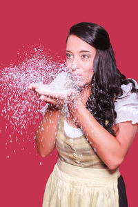 Beautiful young woman blowing artificial snow against red background