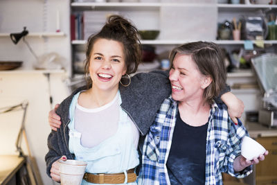 Portrait of happy young potter standing with arm around mature colleague at workshop