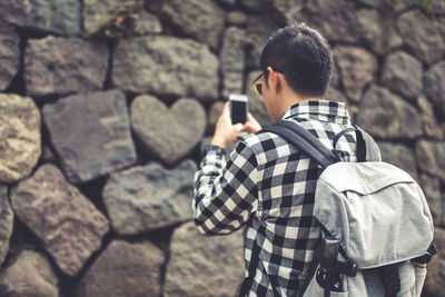 Man wearing backpack while using mobile phone against rock wall