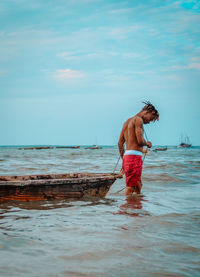 Shirtless man with boat standing at seashore against sky