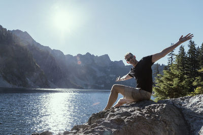 Portrait of mature man with arms outstretched sitting on rock by lake against clear sky