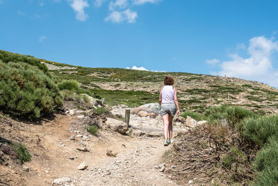 Rear view of woman hiking on dirt road amidst bushes against sky