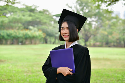 Happy young woman in graduation gown holding folder while standing on field