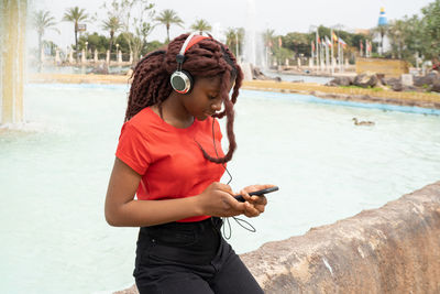 Young focused african american female browsing smartphone while listening to music in headphones near fountain in city