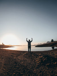 Silhouette man with arms raised standing at lakeshore against sky