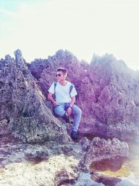 Portrait of young man sitting on rock against sky