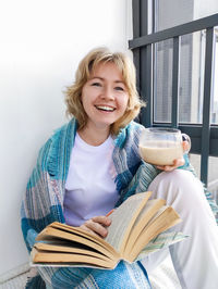 Portrait of a smiling young woman with short blond hair holding a book and coffee cup