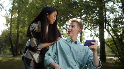 Man sitting on wheelchair holding mobile while talking with girlfriend at park