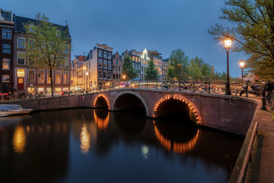 Arch bridge over river amidst buildings against sky at dusk