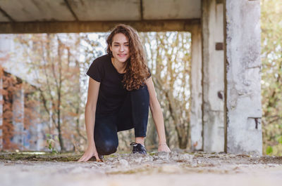 Smiling teenage girl in black jeans squats on abandoned construction. vacation outdoor.