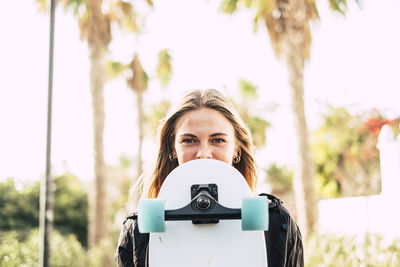 Portrait of woman holding skateboard standing against sky