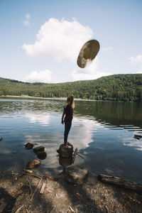 Full length of young woman throwing hat while standing by lake