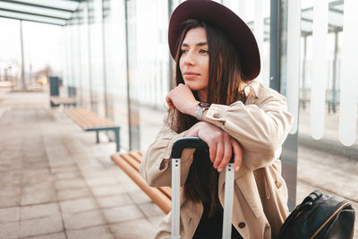 Pensive stylish girl in a coat and hat sits at a stop waiting bus