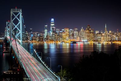 Illuminated bridge over river by buildings against sky at night